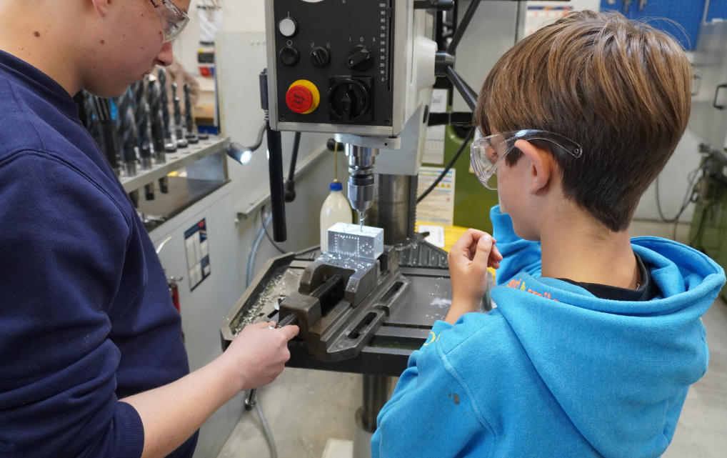 Azubi unterstützt Kind an der Standbohrmaschine in der esmo Werkstatt