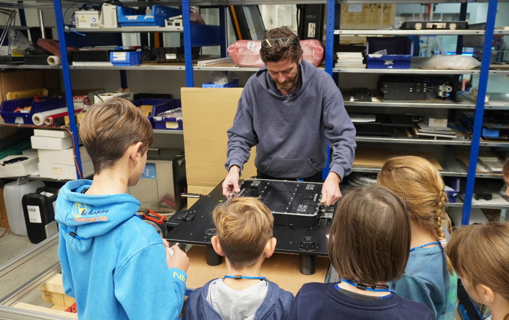 Mitarbeiter erklärt Bauteile auf einer Montageplatte einer Gruppe von Kindern