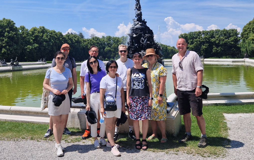 Das internationale Finance-Team der esmo group beim Teamausflug auf der Herreninsel am Chiemsee. Neun Mitarbeiterinnen und Mitarbeiter aus Rosenheim, Shanghai, Singapur und Roseville/Kalifornien stehen lächelnd auf einem Kiesweg in sommerlicher Kleidung. Im Hintergrund grüne Wiesen, Bäume und blauer Himmel mit weißen Wolken.