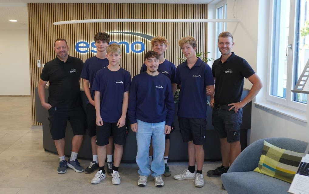 Five new esmo AG apprentices stand with two supervisors in the entrance area at the Rosenheim site.