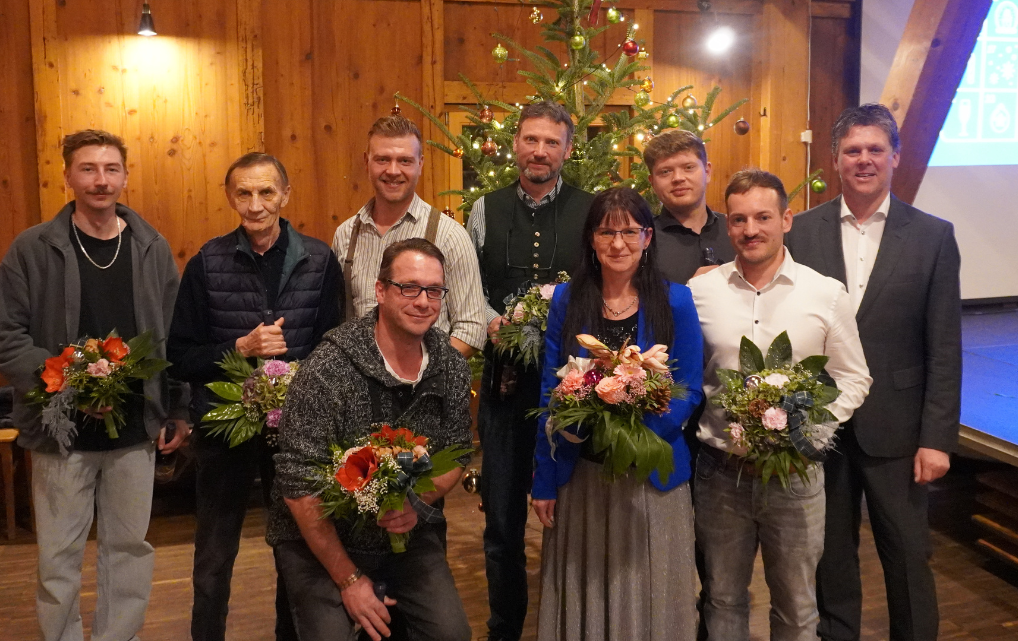 Group photo from the esmo Christmas celebration 2025: Several employees stand in a festively decorated hall in front of a decorated Christmas tree, holding flower bouquets in recognition of their 10- and 20-year company anniversaries. The scene reflects a warm and appreciative atmosphere.